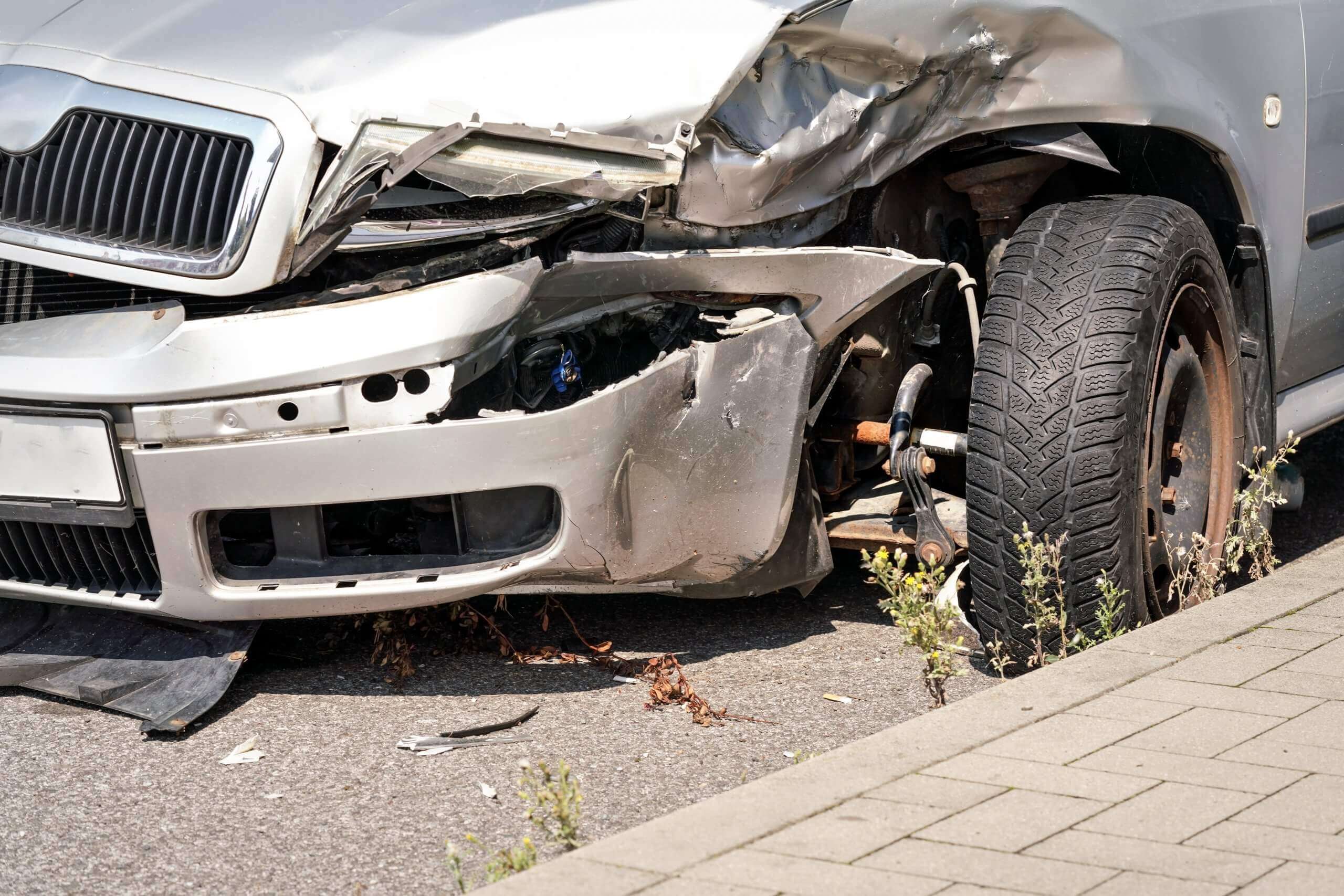 Die stark beschädigte Front eines silbernen Autos nach einem Unfall mit zerbeulter Stoßstange und freiliegendem Rad, relevant für ein Schadengutachten