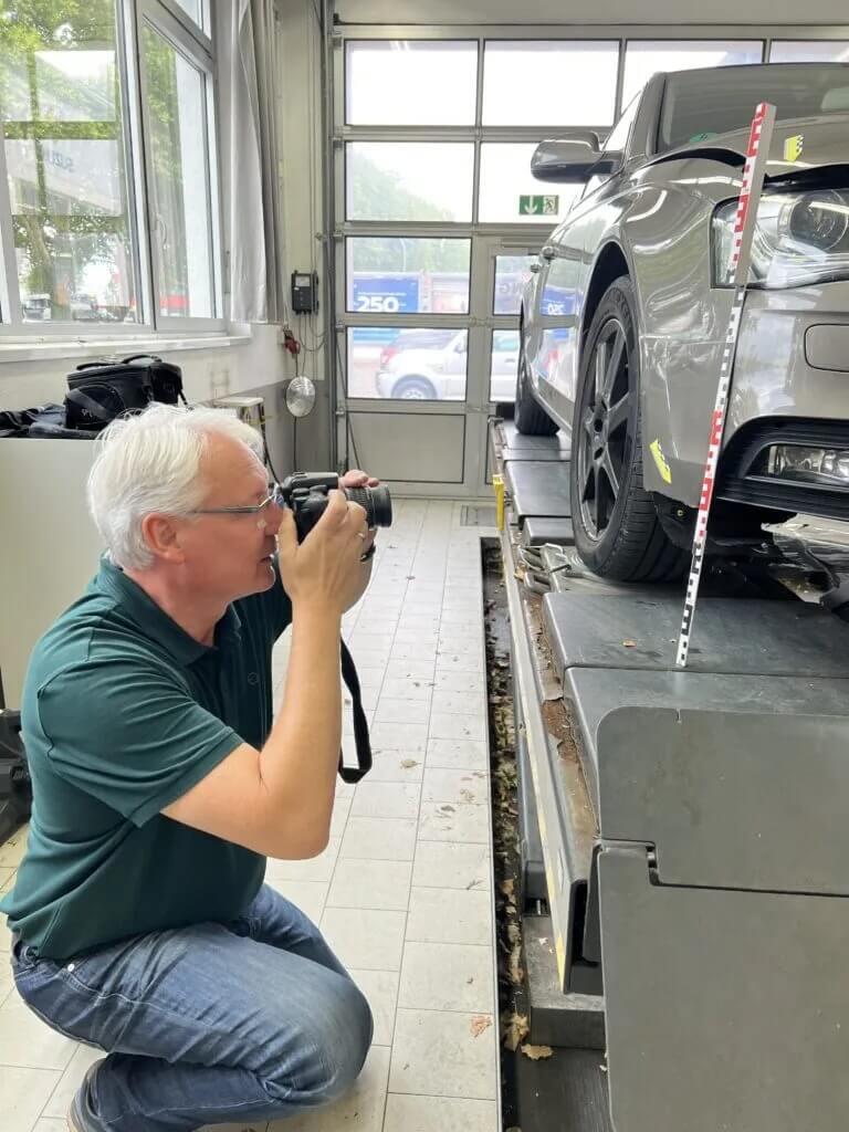 Ein Mann kniet in einer Garage und fotografiert die graue Stoßstange eines Autos, neben dessen Rad ein Messstab steht