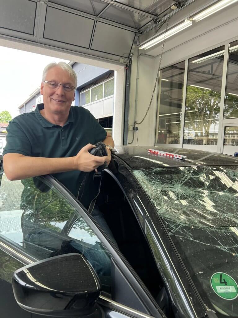 Ein Mann steht in einer Werkstatt neben einem Auto mit zerbrochener Windschutzscheibe und lächelt