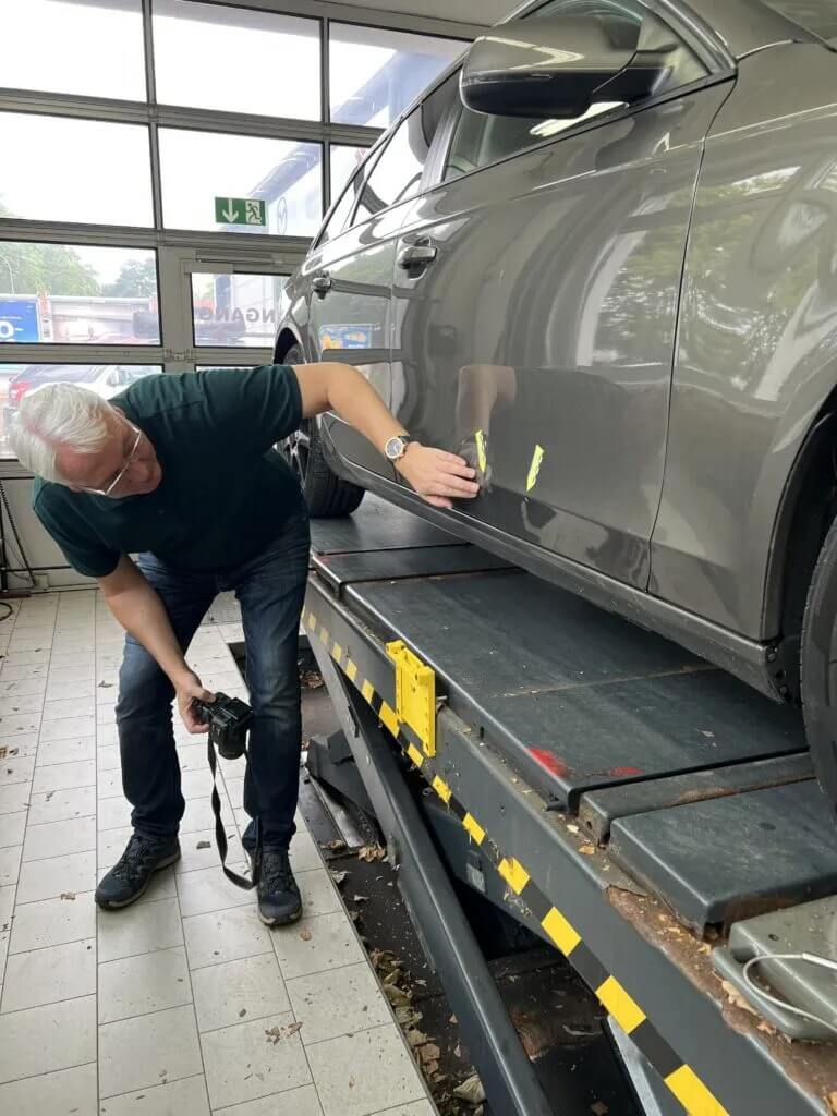 Ein Mann untersucht und fotografiert Kratzer an der Seite eines grauen Autos, das auf einer Hebebühne in einer Werkstatt steht