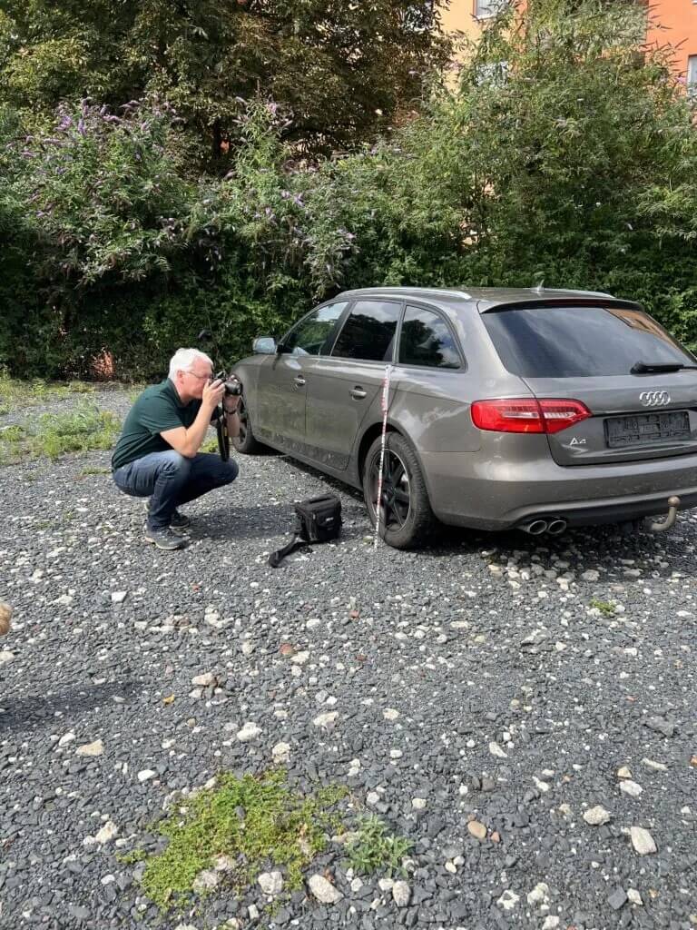 Ein Mann kniet und fotografiert die Seite eines staubgrauen Audi-Kombis auf Schotter, neben dem Auto steht eine Messlatte
