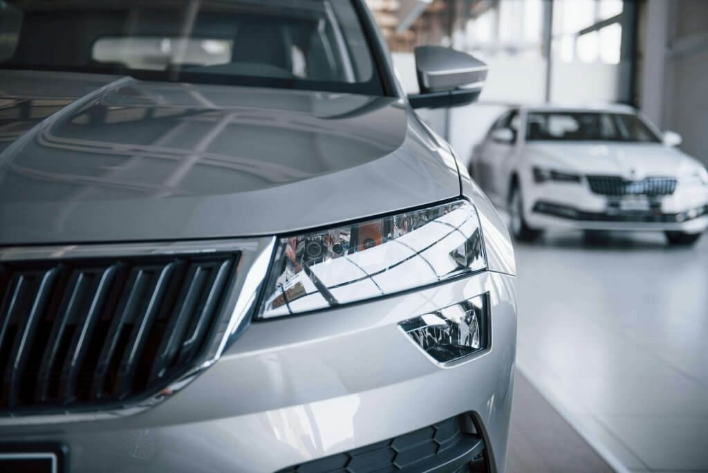 Nahaufnahme von Frontscheinwerfer und Kühlergrill eines silbernen Autos im hellen Showroom, geeignet für Schadengutachten, dahinter weiteres Auto