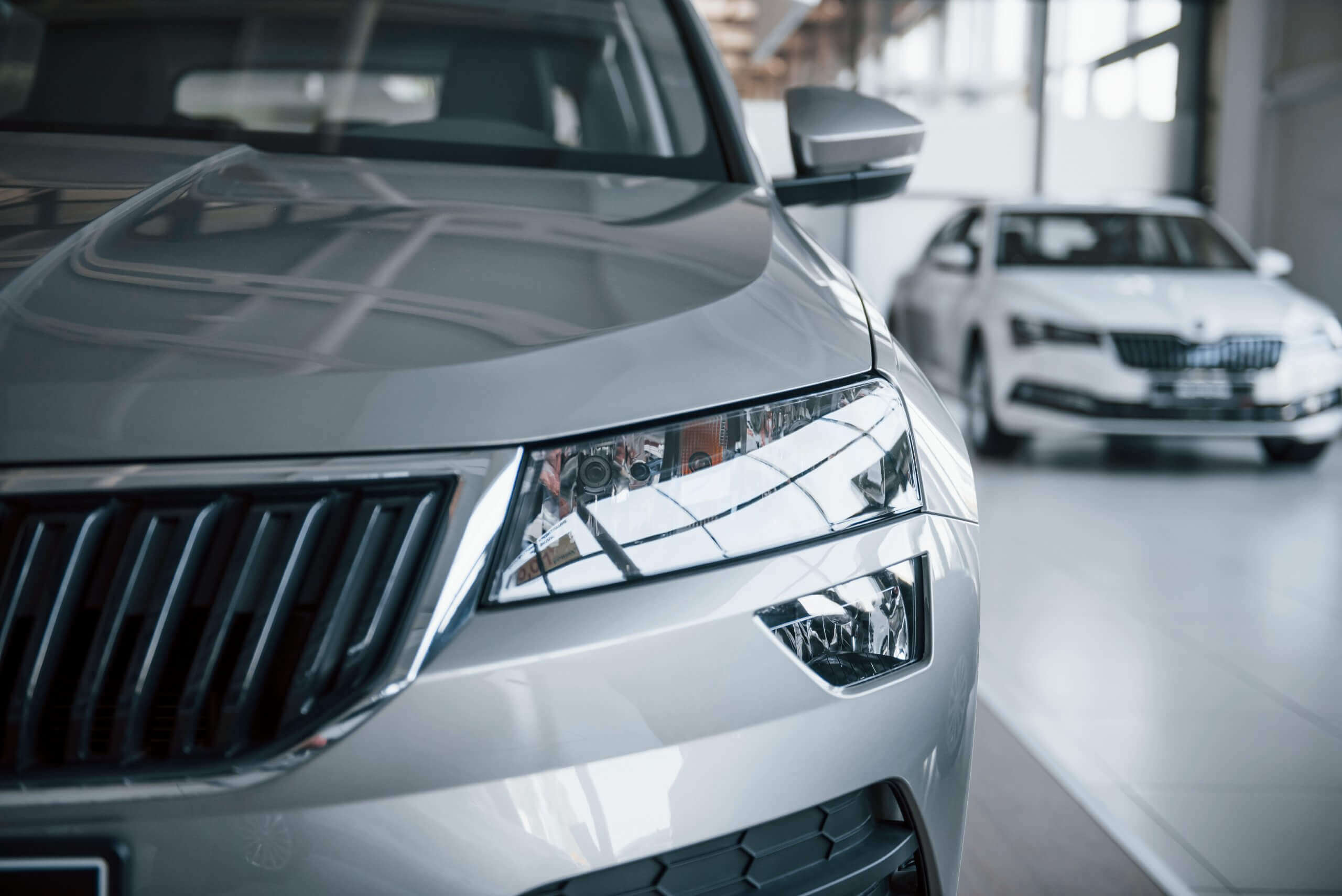 Nahaufnahme von Frontscheinwerfer und Kühlergrill eines silbernen Autos im hellen Showroom, geeignet für Schadengutachten, dahinter weiteres Auto