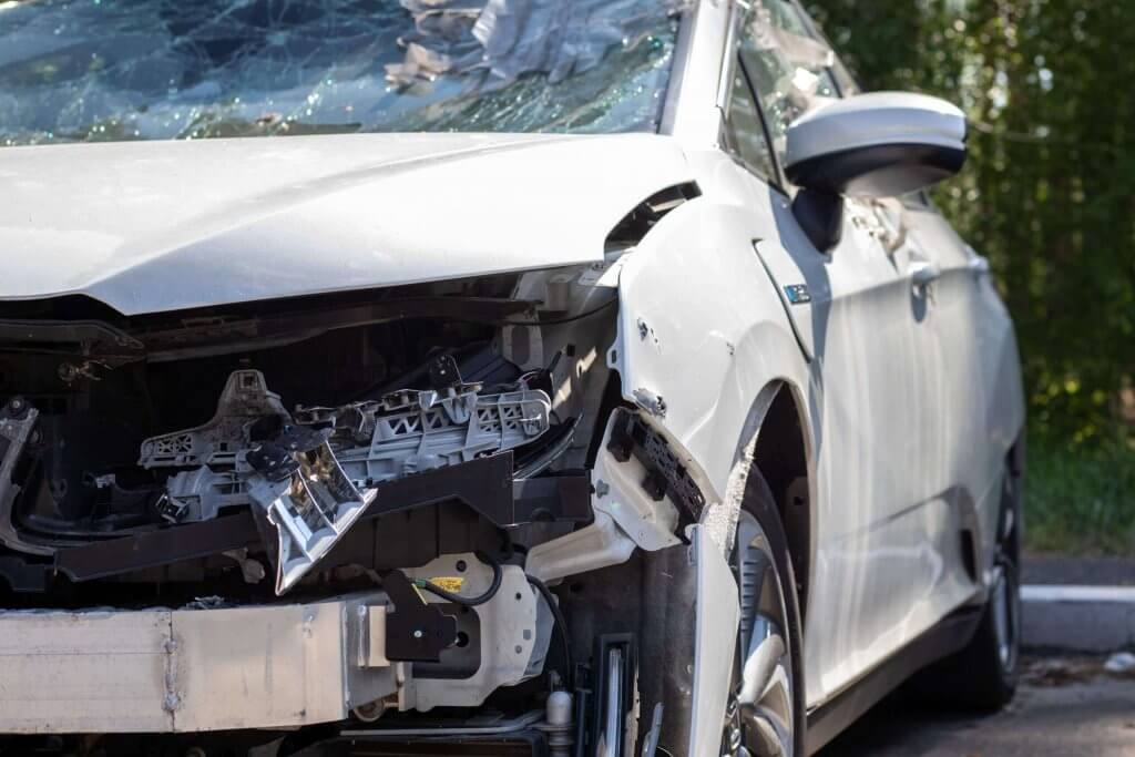 Ein weißes Auto mit stark beschädigter Front und zersplitterter Windschutzscheibe steht tagsüber draußen, geeignet für ein Schadengutachten