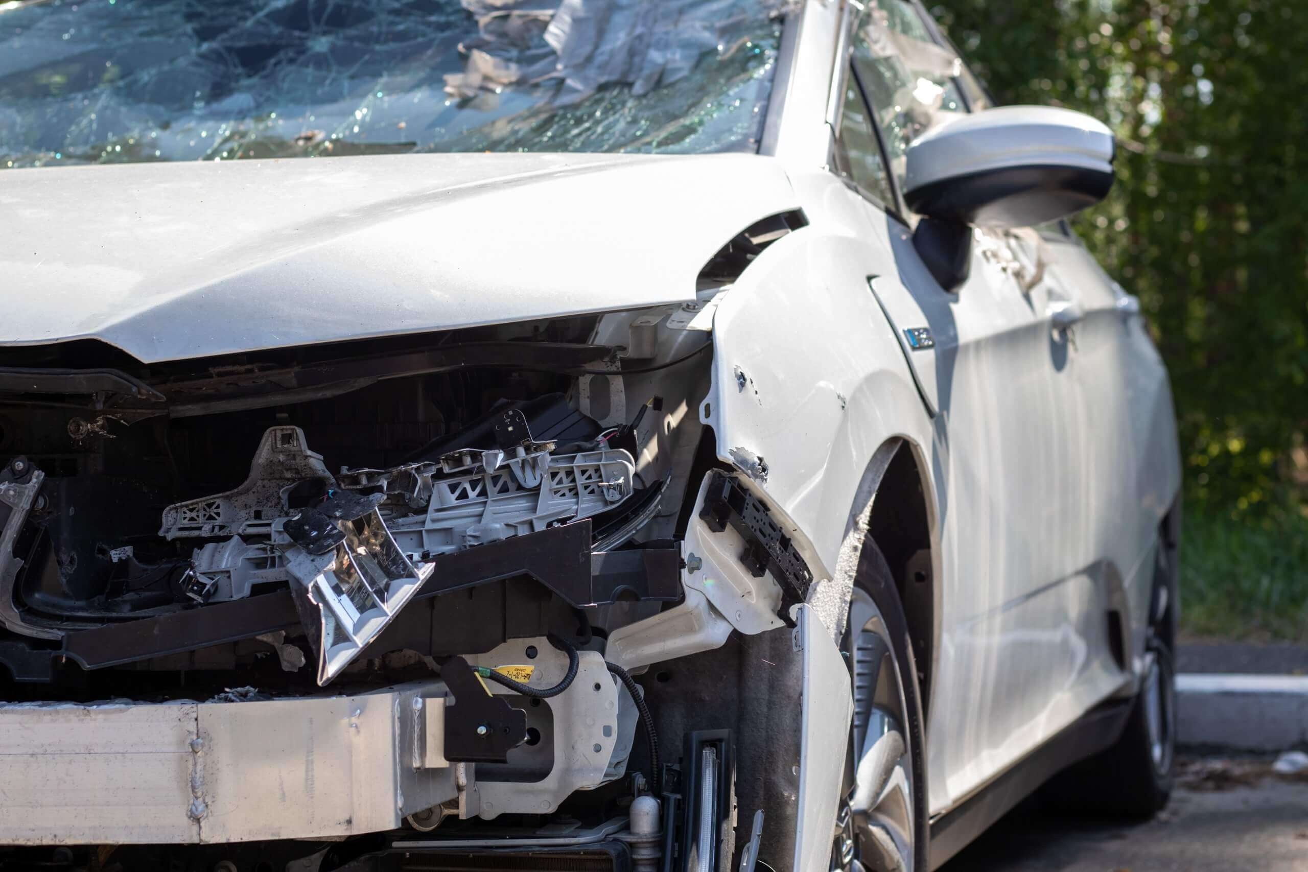 Ein weißes Auto mit stark beschädigter Front und zersplitterter Windschutzscheibe steht tagsüber draußen, geeignet für ein Schadengutachten