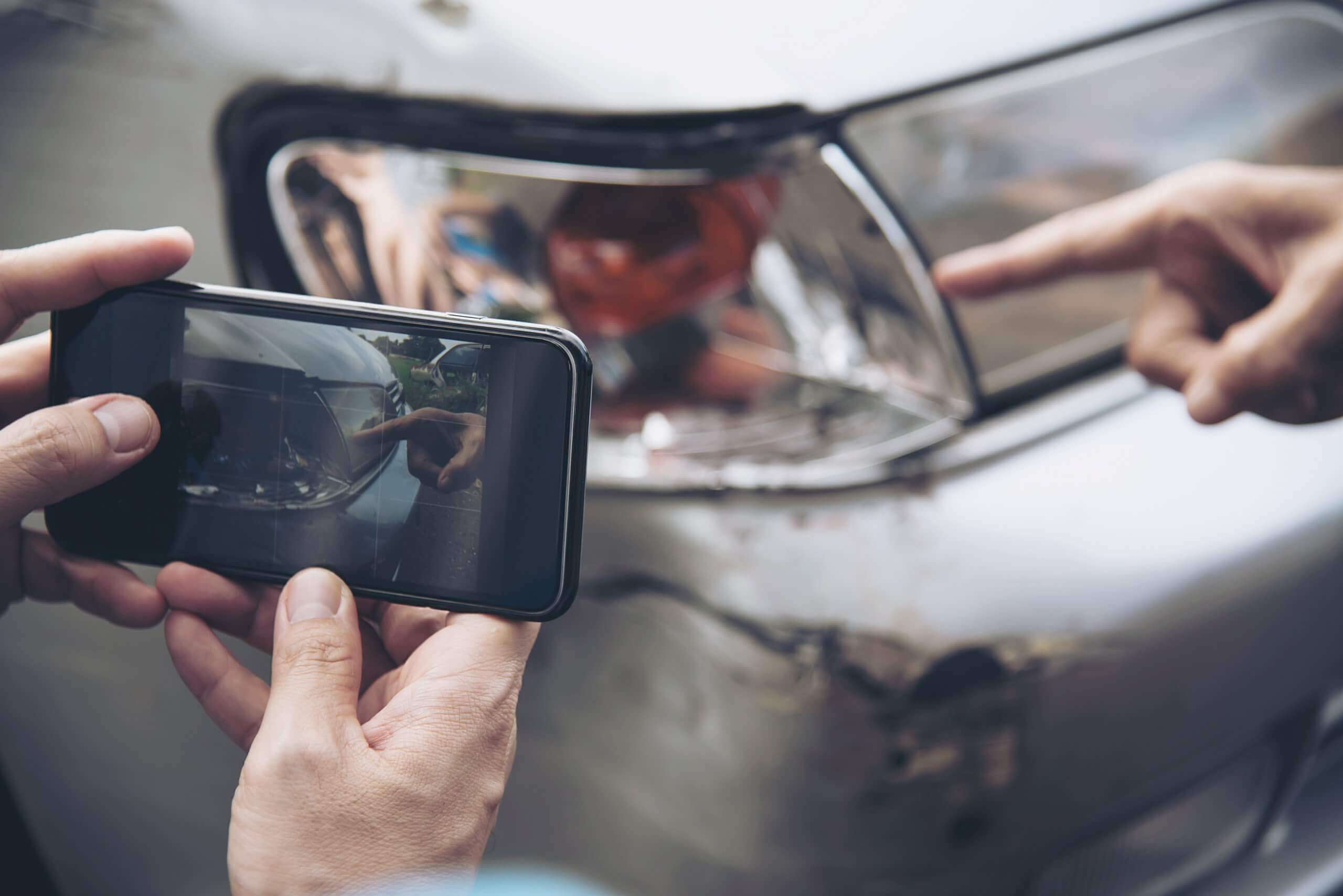 Eine Person fotografiert für ein Schadengutachten einen Blechschaden am Auto, während jemand auf die Delle neben dem Scheinwerfer zeigt