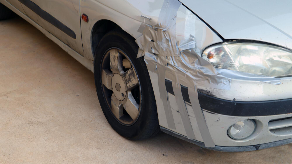 Ein silbernes Auto mit Frontschaden, dessen Stoßstange provisorisch mit grauem Klebeband fixiert ist und wie ein Entwurf wirkt